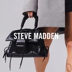 Mujer con blazer de cuadros de gran tamaño sobre top corto negro y shorts, sosteniendo un bolso negro, de pie en una habitación blanca con tragaluz.