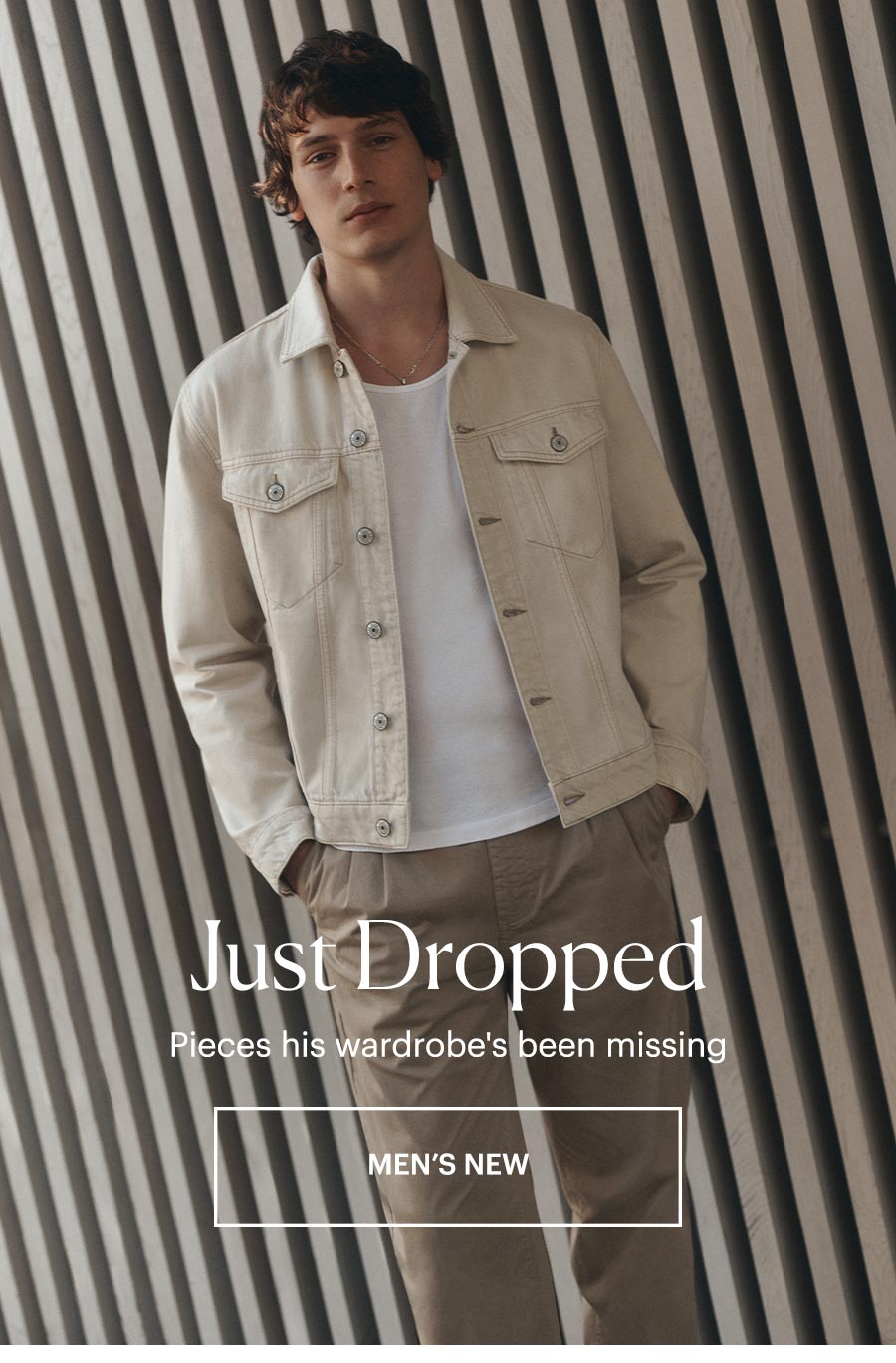 Man wearing a cream denim jacket over a white tee with tan trousers, standing against a white wood slat wall. Just Dropped - Pieces his wardrobe's been missing. SHOP MEN'S NEW ARRIVALS