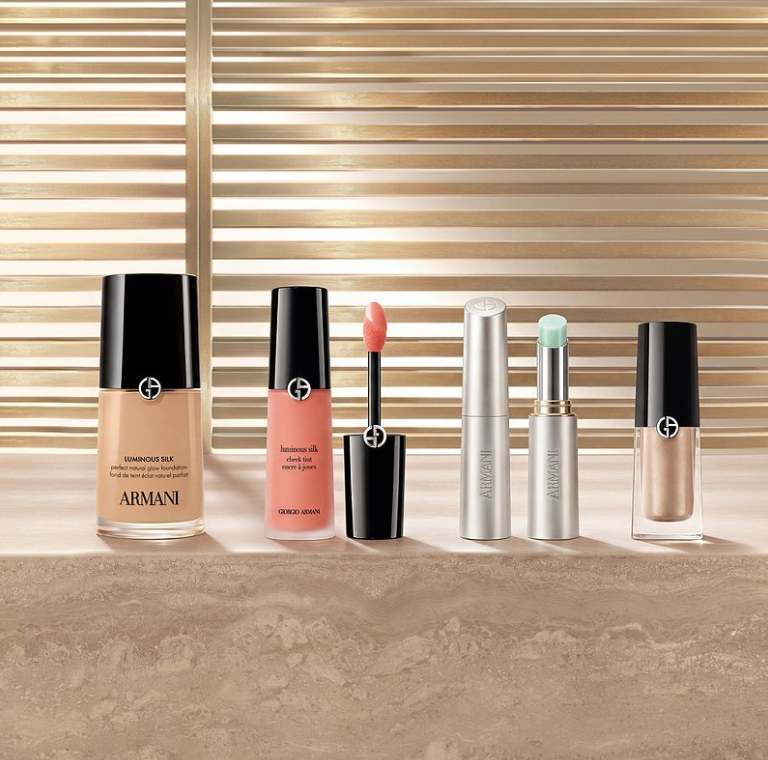 Los productos para el rostro de Armani beauty están presentados en fila sobre una superficie de piedra, delante de un fondo de lamas en tonos dorados. La selección incluye una base Luminous Silk, un tinte rosa para mejillas con su aplicador abierto, dos sticks plateados de cuidado o de tez y un producto líquido luminoso.