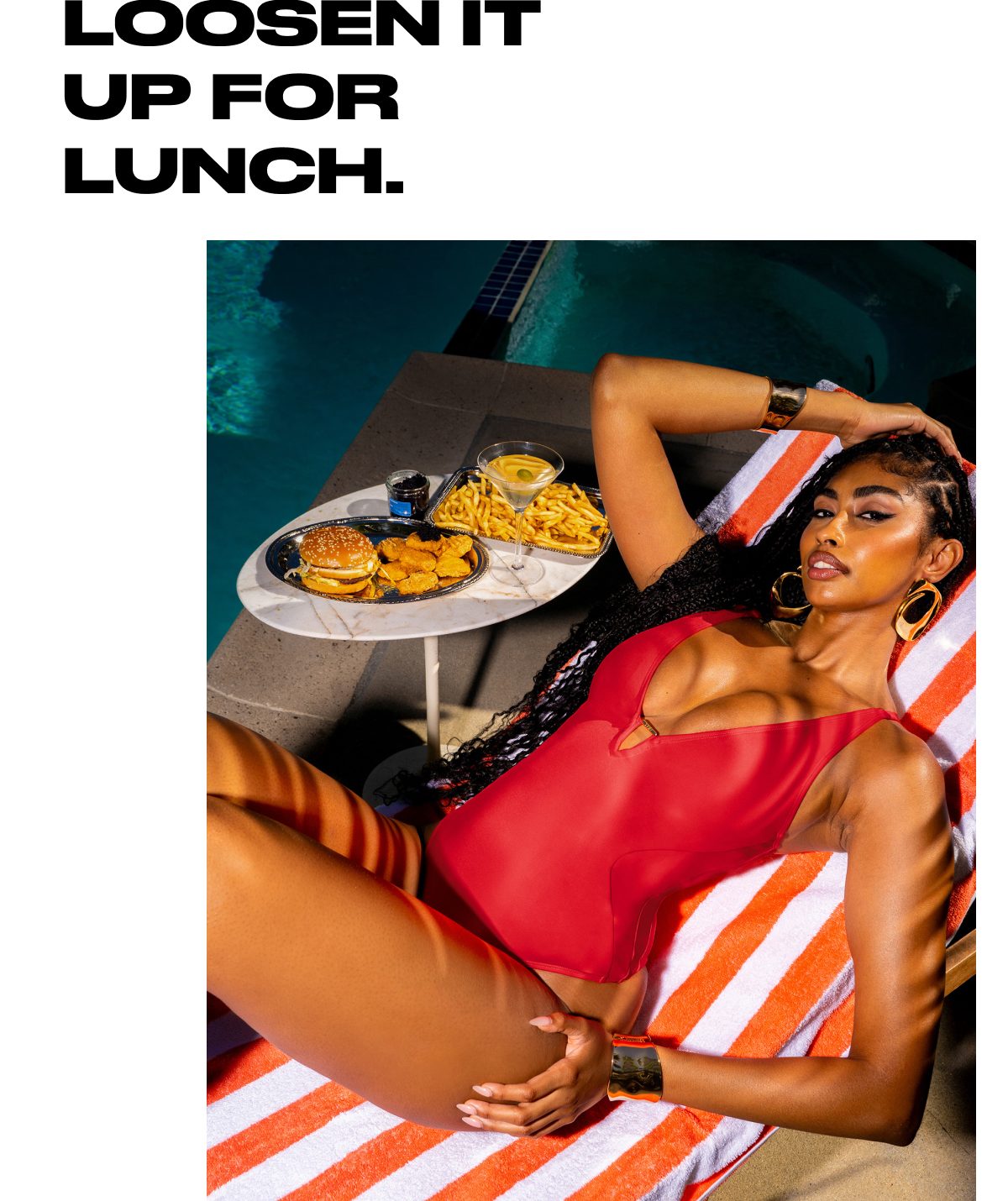 A woman in a red swimsuit relaxes by a pool with lunch on a table beside her.