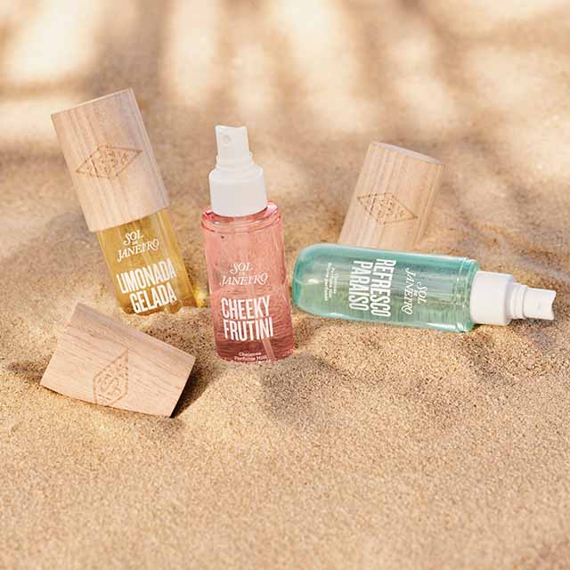 Three Sol de Janeiro perfume bottles are displayed on sand, with wooden caps beside them.