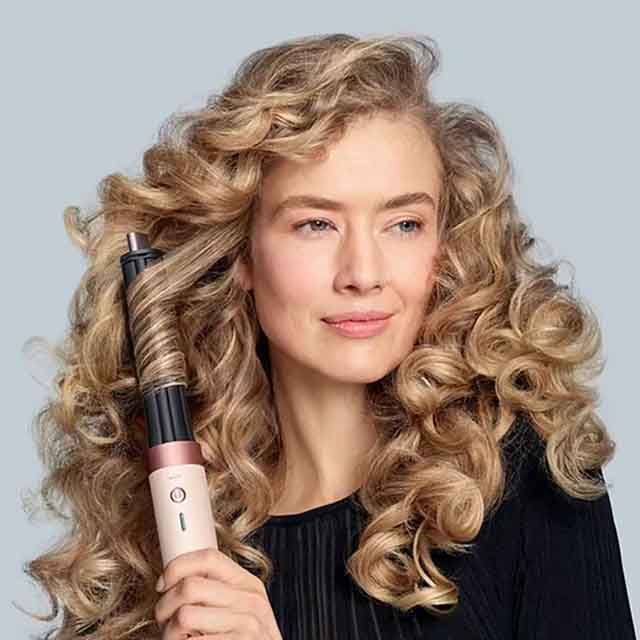 A person with curly hair holds a curling iron, showcasing styled hair.