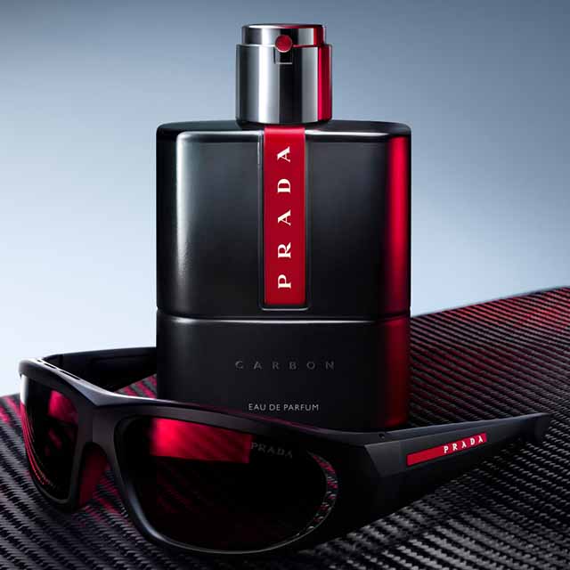 A black Prada perfume bottle beside black sunglasses with red accents on a textured surface.