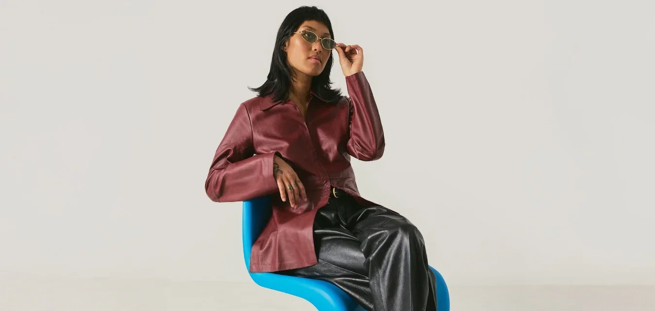 Vrouw met een bordeauxrode jas en zwarte broek zit op een moderne blauwe stoel en stelt haar zonnebril af tegen een effen lichte achtergrond.