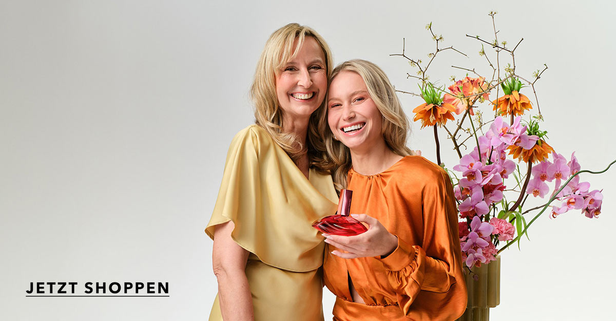 Zwei lächelnde Frauen stehen eng beieinander vor einem hellen Hintergrund. Die ältere Frau trägt ein goldenes Kleid, die jüngere ein orangefarbenes Kleid und hält ein rotes Parfümfläschchen in der Hand. Neben ihnen steht eine Vase mit bunten Blumen. Unten links steht der Schriftzug „JETZT SHOPPEN“.
