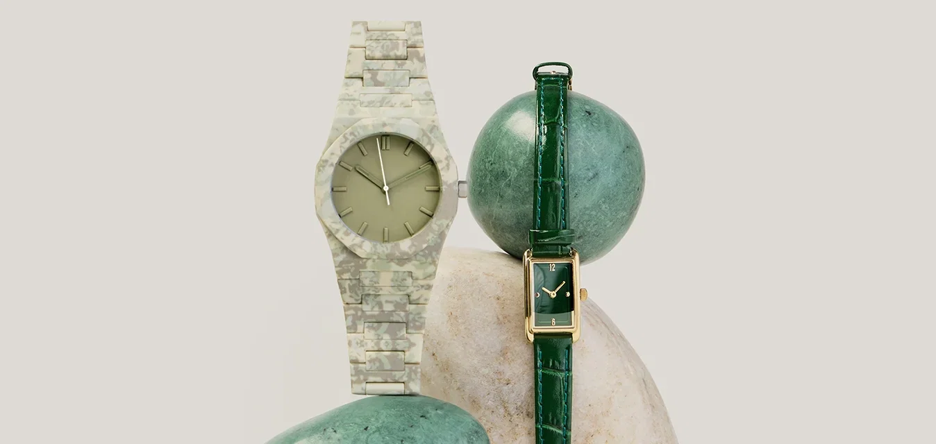 Dos relojes de pulsera, uno con correa de cuero verde y esfera rectangular, el otro con banda de patrón camuflaje y esfera redonda, exhibidos sobre piedras.