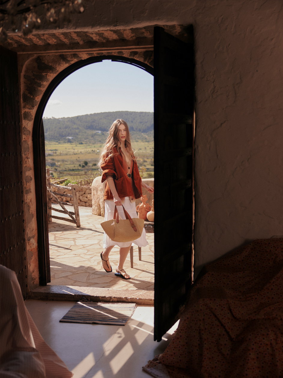 Ein Model von DOROTHEE SCHUMACHER steht in einem rustikalen Torbogen mit Blick auf eine sonnige mediterrane Hügellandschaft. Sie trägt eine warme, rostfarbene Jacke über einem luftigen weißen Rock, flache Sandalen und hält eine große Strohtasche in der Hand – ein Look, der sommerliche Eleganz und Reiselust perfekt vereint.