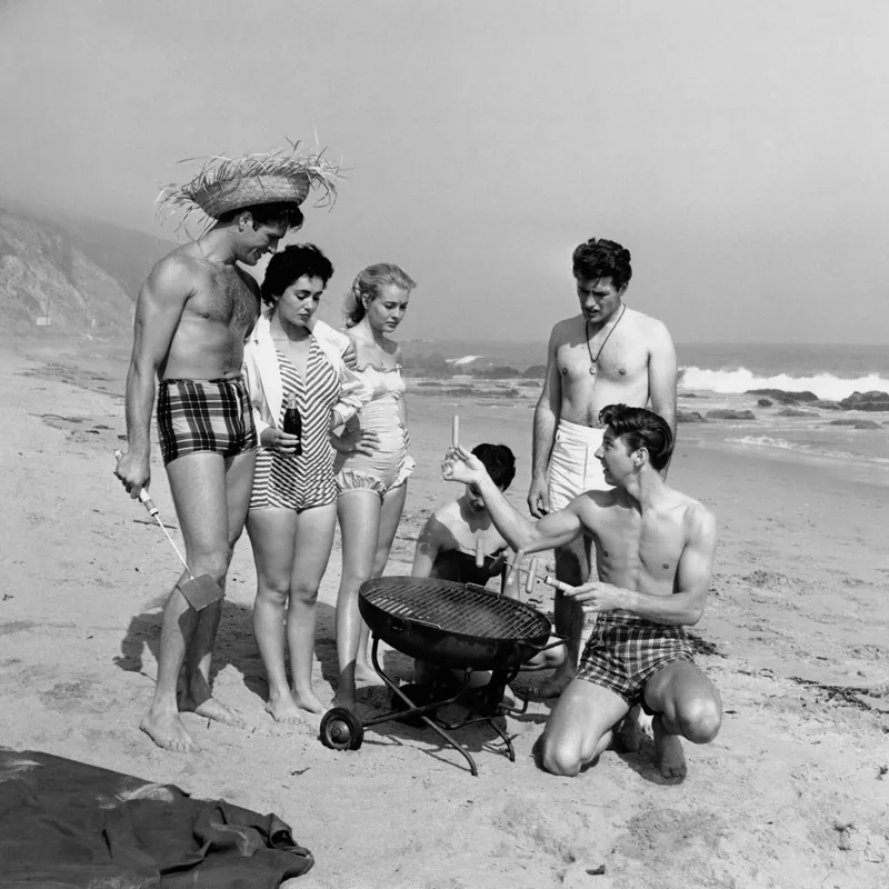 American actor Hugh O'Brian (1925 - 2016, standing, right) enjoys a barbecue on the beach with friends, circa 1955.