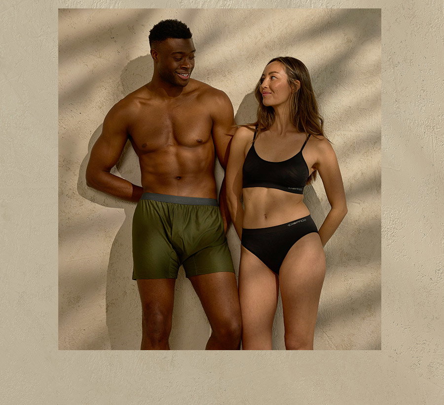 A man in green boxer shorts and a woman in a black bra and underwear standing together against a textured wall.