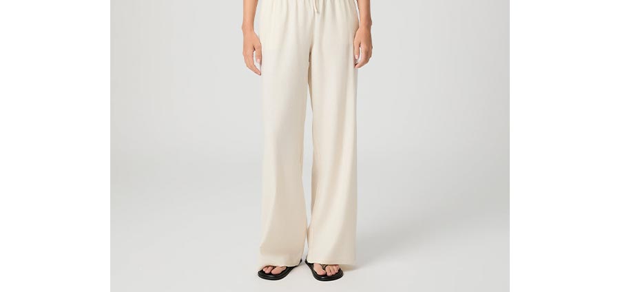 Woman wearing cream linen wide leg pants and black flip flop sandals in front of a grey backdrop.