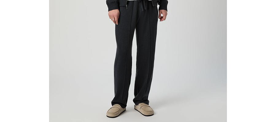 Man wearing a pair of deep charcoal grey lounge pants with beige suede shearling slides in front of a grey backdrop.