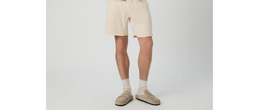 Man wearing a pair of cream drawstring lounge shorts with white socks and white suede shearling slides in front of a grey backdrop.