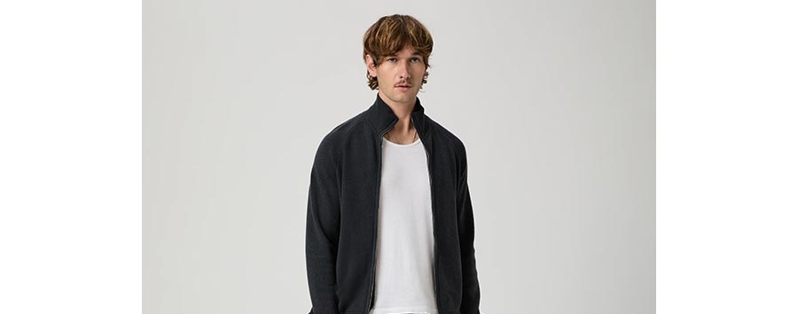 Man wearing a dark grey zip-up sweater over a white tee in front of a grey backdrop.