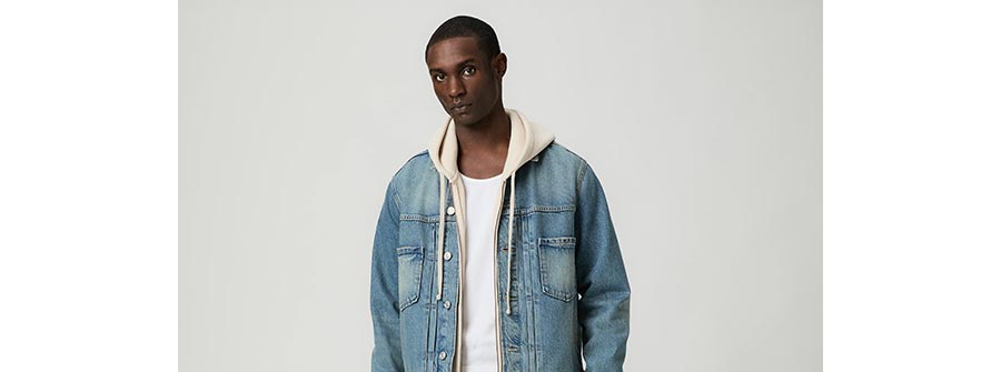 Man wearing a light wash classic denim jacket over a cream zip-up hoodie and white tee, in front of a grey backdrop.