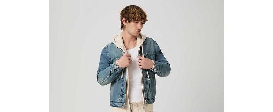 Man wearing a light wash classic denim jacket over a cream zip up hoodie and white tank in front of a grey backdrop.