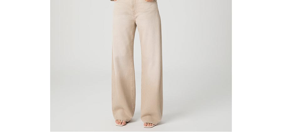 Woman wearing a pair of beige high-rise barrel leg jeans with white flip flop sandal heels in front of a grey backdrop.