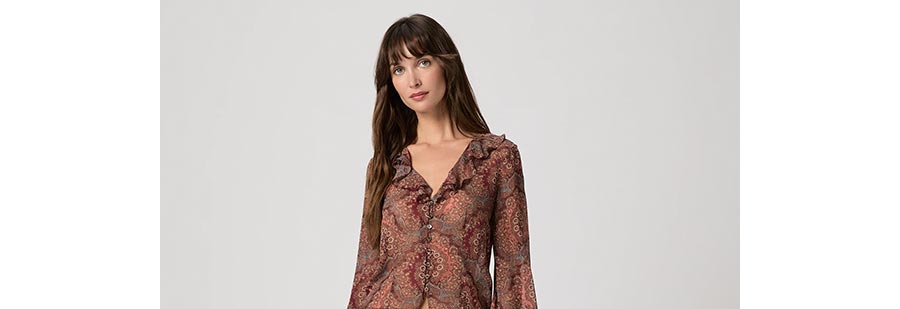 Woman wearing a silk medallion print blouse with asymmetrical flare sleeves, button-front closure, delicate ruffle trim, and a V-neckline.