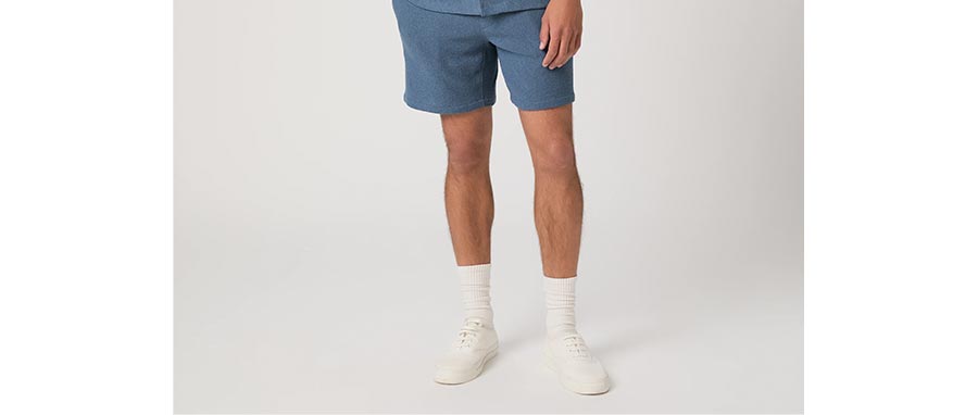 Man wearing a pair of blue micro boucle cotton blend shorts with white socks and sneakers, standing against a grey backdrop.