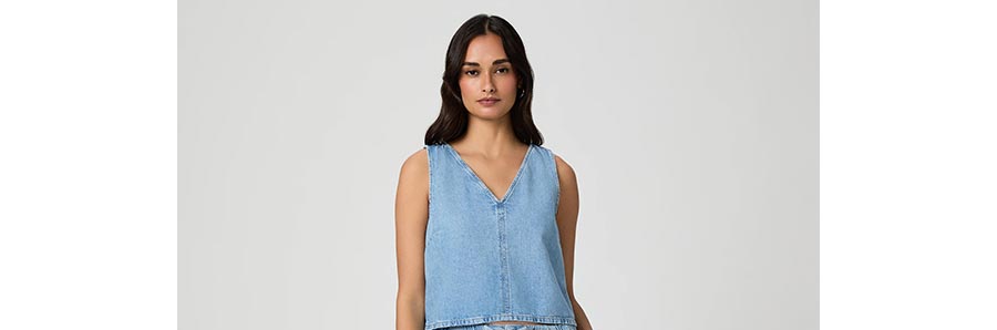 Woman wearing a cropped, boxy fit blue denim tank top in front of a grey backdrop.