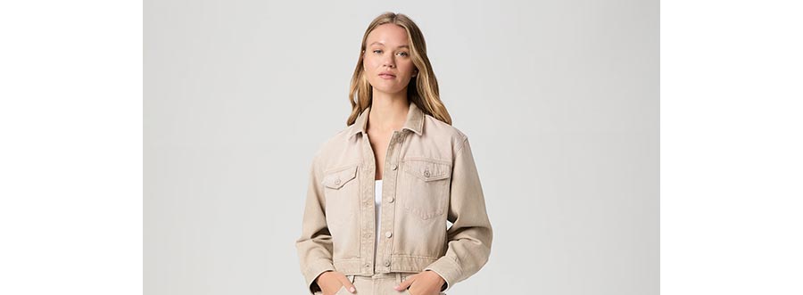Woman wearing a cropped beige denim jacket over a white tank in front of a grey backdrop.