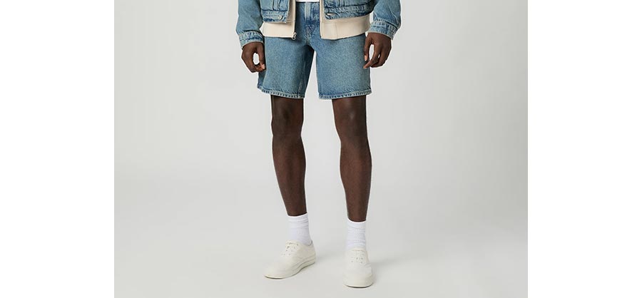 Man wearing a pair of light wash denim shorts with white socks and sneakers in front of a grey backdrop.
