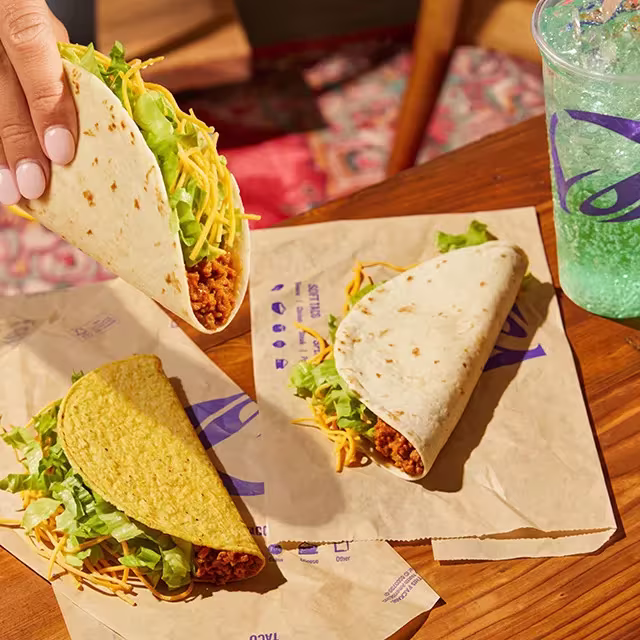 A hand holding a soft taco next to a soft drink, a crunchy taco, and another soft taco on a table.