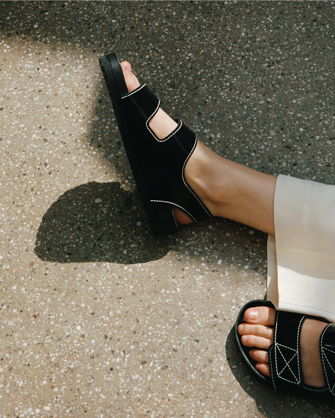 Cropped image of models legs laying on pavement, wearing the Birkenstock x Proenza Schouler Milano Sandals in Black with the Nila Pant in Cotton Drill in Marzipan