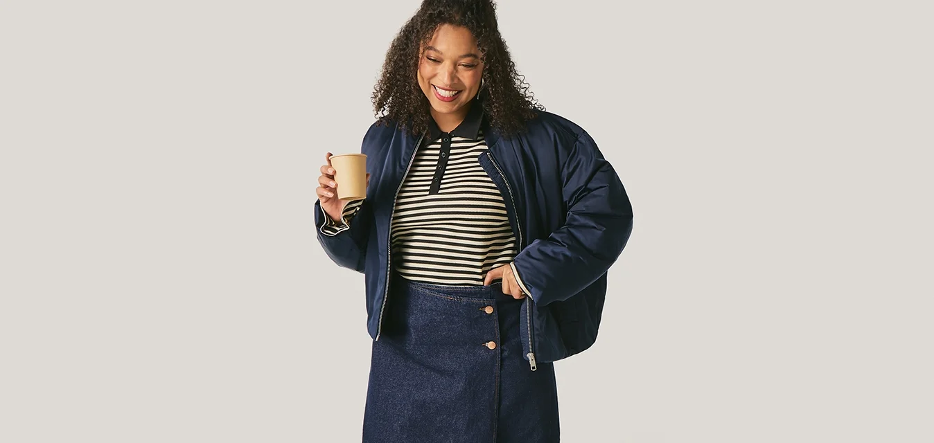 Lachende vrouw in een marineblauwe jas, gestreept shirt en rok houdt een koffiekop vast, staand tegen een effen lichte achtergrond.