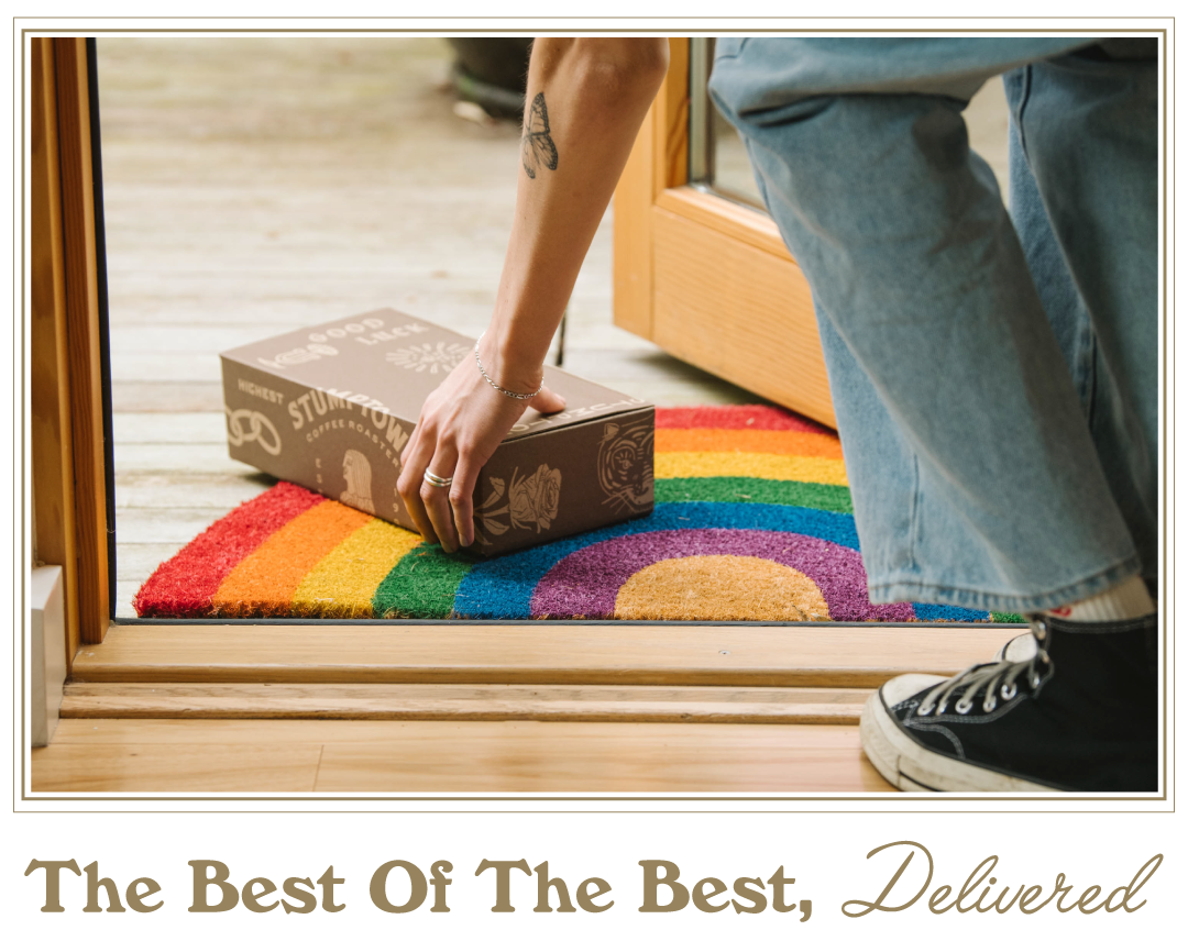 A person picks up a delivered Stumptown Roaster’s Pick subscription coffee box from a rainbow doormat at a front doorstep, with the headline “The Best Of The Best, Delivered.”