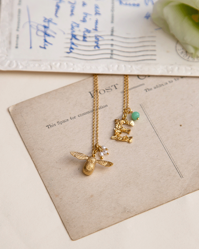 Gold bee and initial necklaces with gemstone detail styled on vintage postcards with a pen and soft floral touch