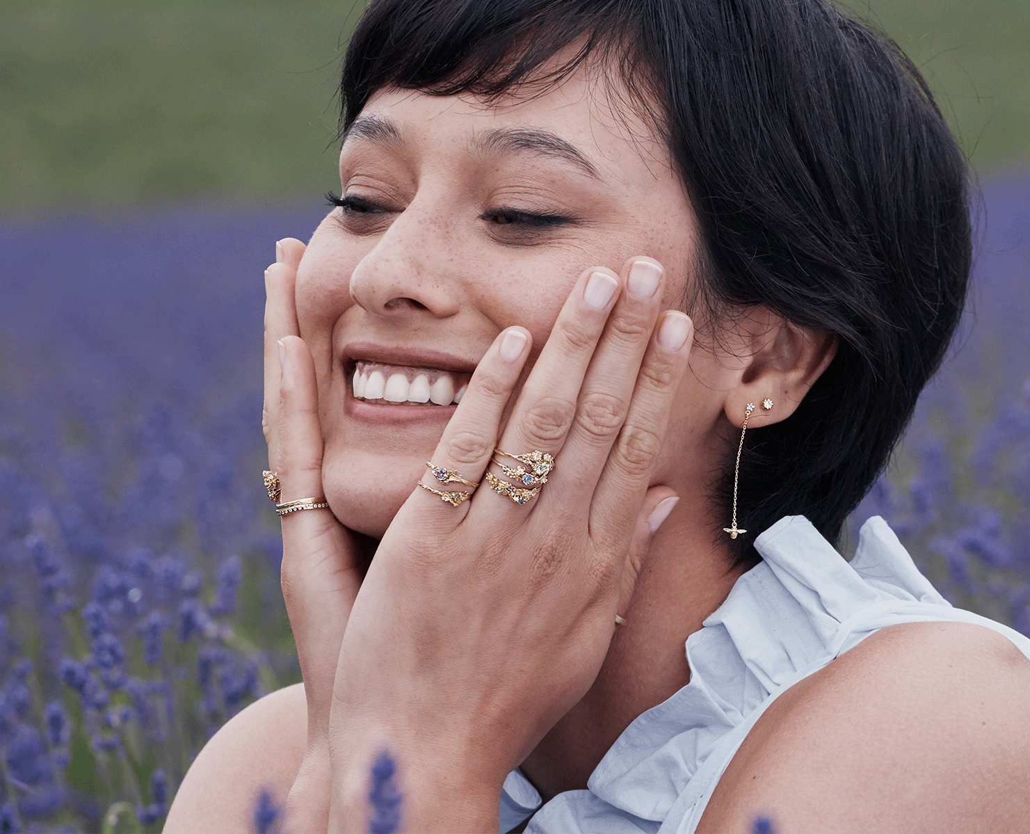 Woman wearing multiple gold rings with colourful gemstones and delicate drop earrings, styled in a lavender field with soft natural light.