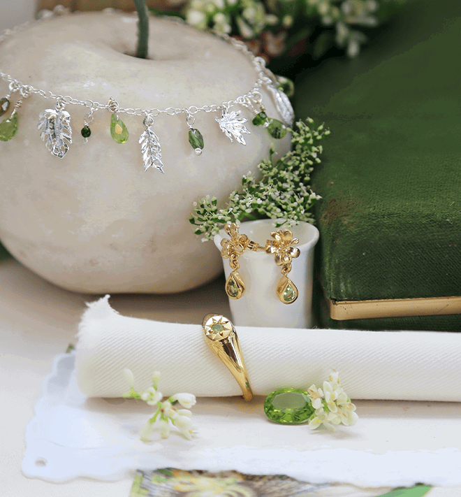 Silver leaf charm bracelet with green gemstone drops, styled alongside gold floral drop earrings and a gold ring, set with green stones against soft natural textures and foliage.