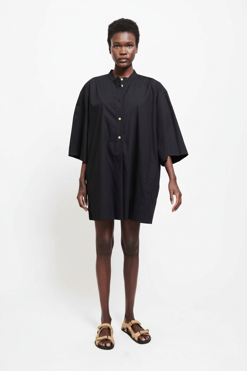 Woman in a black oversized shirt dress and tan sandals stands against a white background.