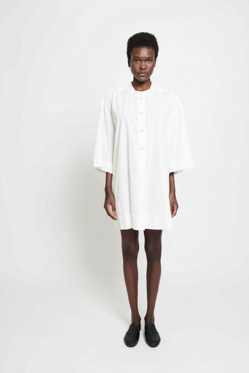 A woman in a white shirt dress and black shoes stands against a white background.