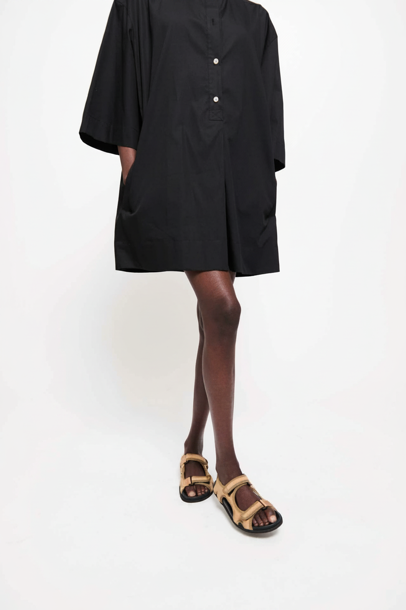 A person wearing a black dress and tan sandals stands against a white background.