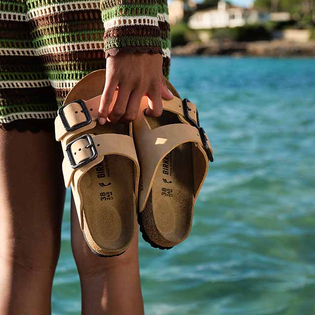 A person in a striped dress holds beige sandals by clear blue water.