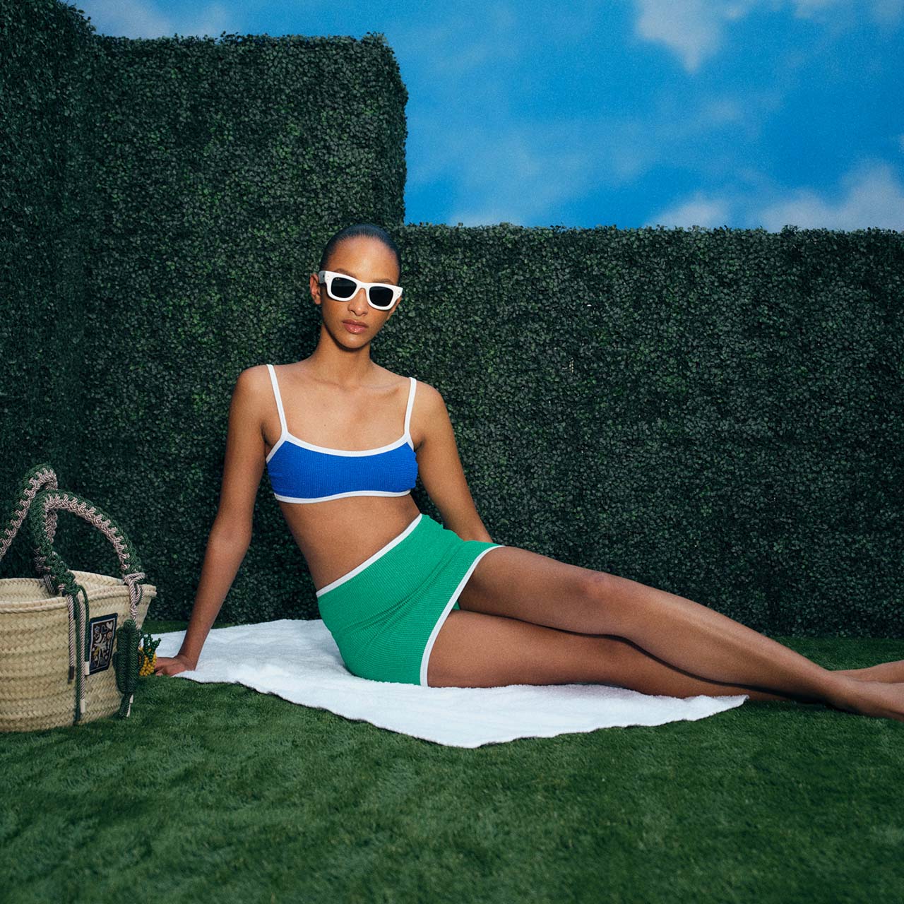 A person in a blue and green outfit, wearing sunglasses, sits on grass against a hedge backdrop with a straw bag nearby.