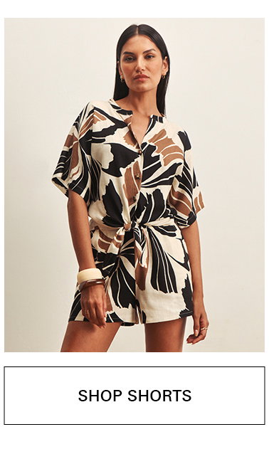 Woman wearing a matching short-sleeve button-up top and shorts with a bold black, brown, and cream abstract print, standing confidently. Text below reads "SHOP SHORTS.