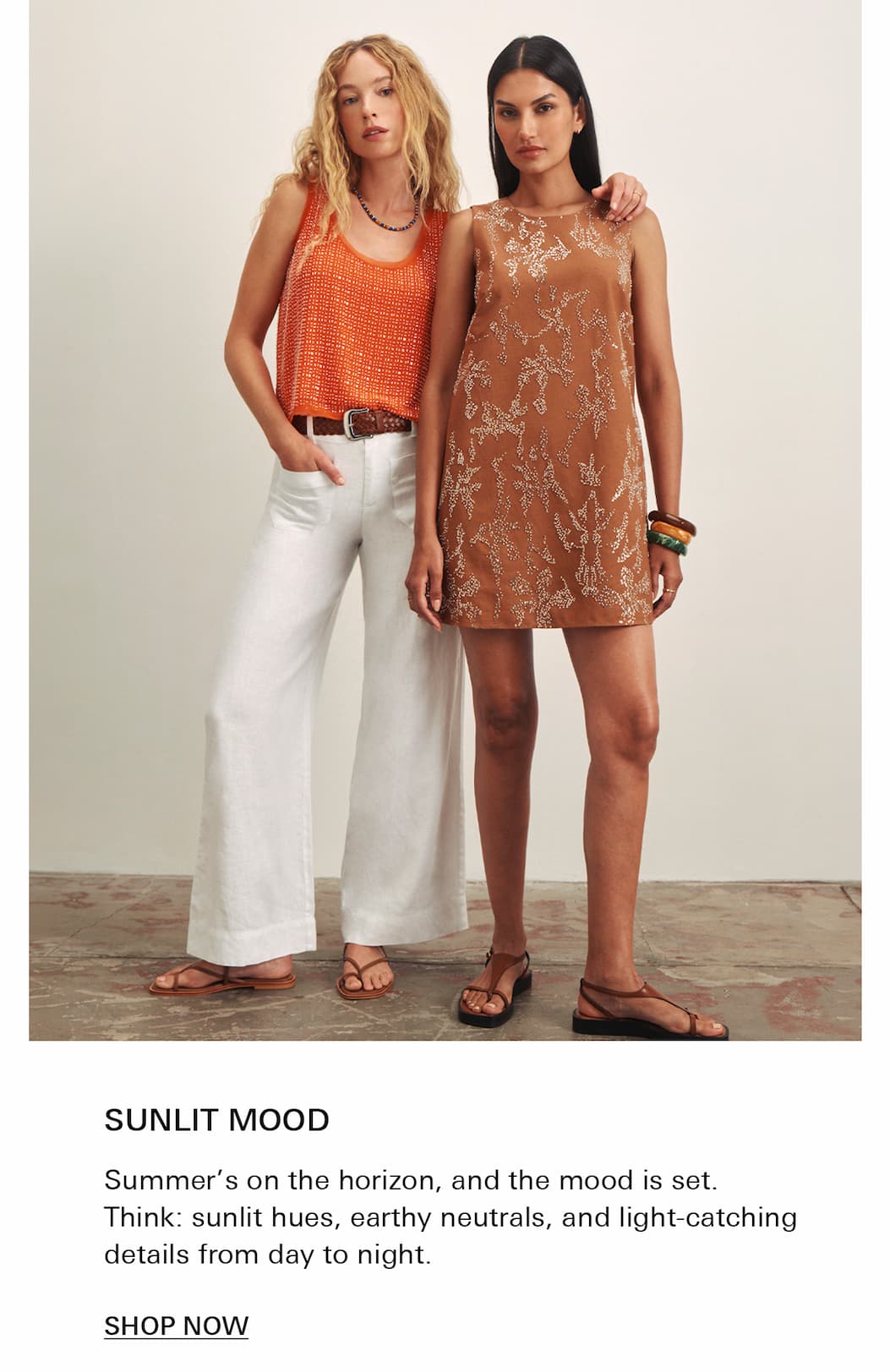 Two women pose indoors against a plain wall. One wears a sleeveless orange top with white wide-leg pants; the other wears a sleeveless tan dress with a delicate white pattern. Both wear sandals and relaxed, summery expressions.