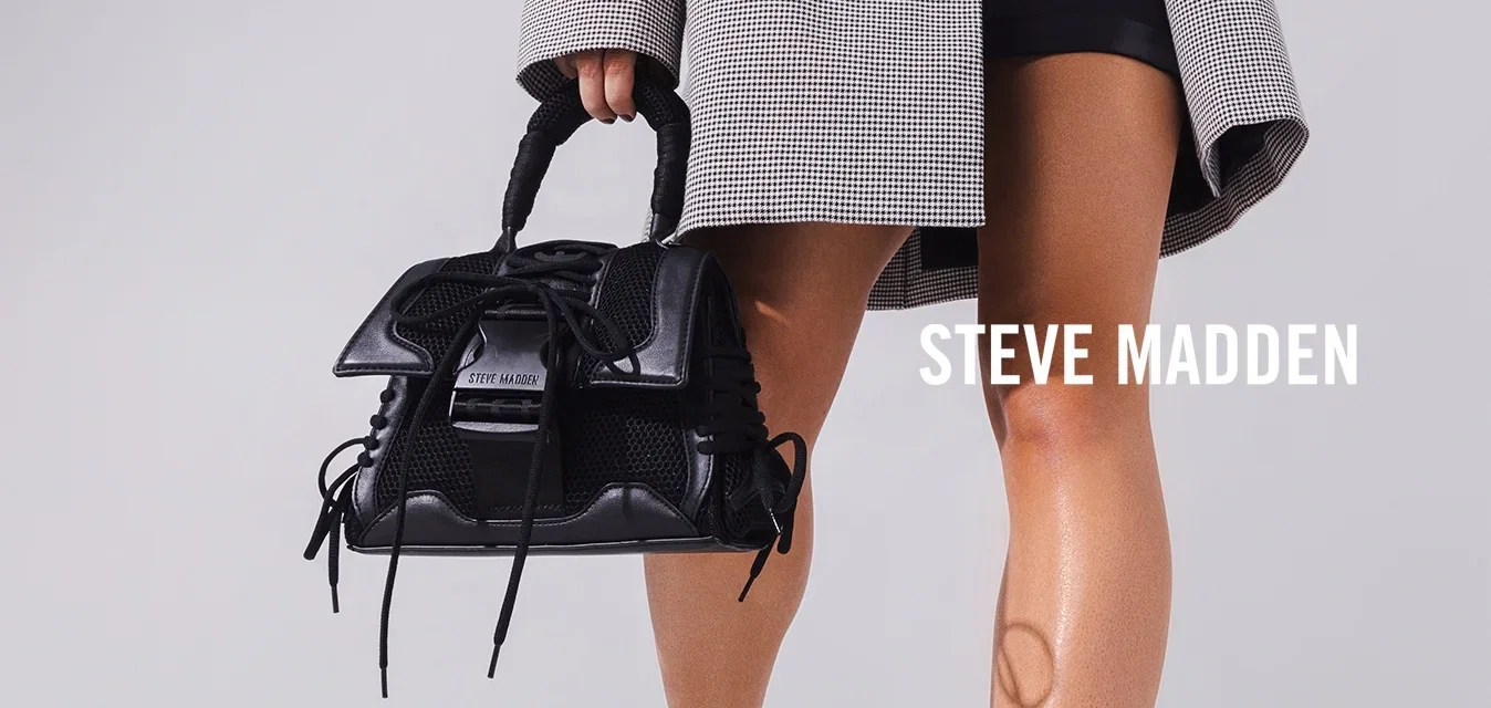 Femme portant un blazer à carreaux oversize par-dessus un crop top noir et un short, tenant un sac à main noir, debout dans une pièce blanche avec un puits de lumière.