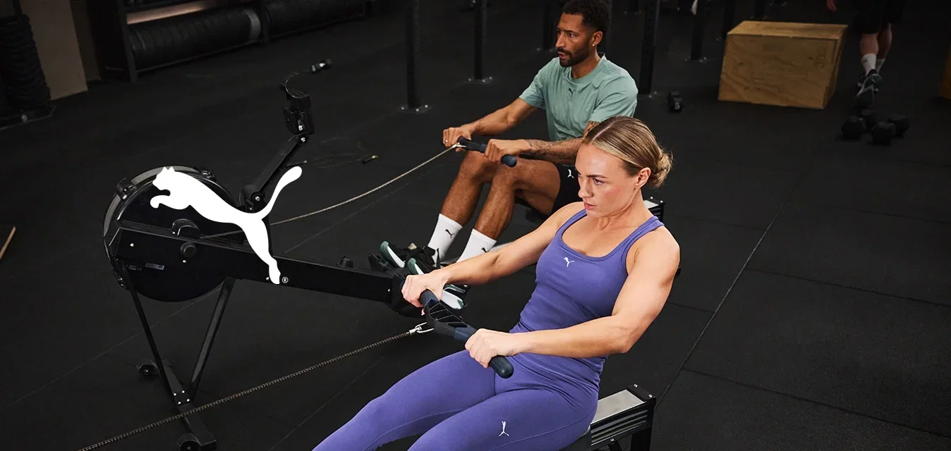 Máquina de remo situada en un gimnasio. Dos usuarios con ropa deportiva; uno con leggings morados y top, y el otro con una camiseta verde. Suelo y equipo de color negro.