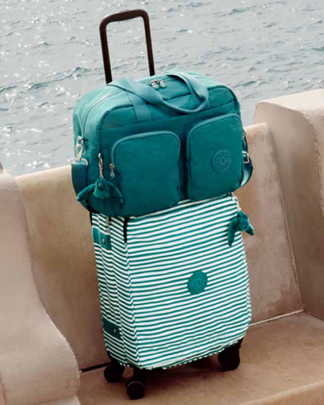 A blue Kipling duffel bag sits on top of a striped suitcase near a body of water.