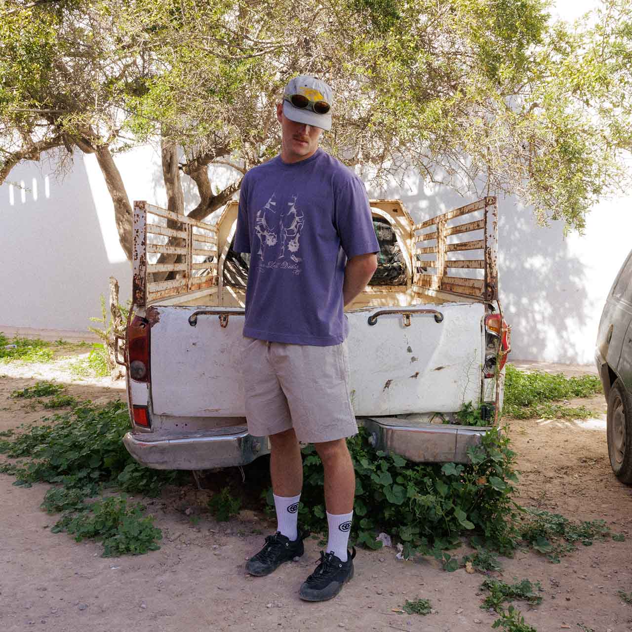 A person in Emporium brand T-shirt, shorts and branded socks, standing in front of an old truck bed, with trees and a wall in the background.