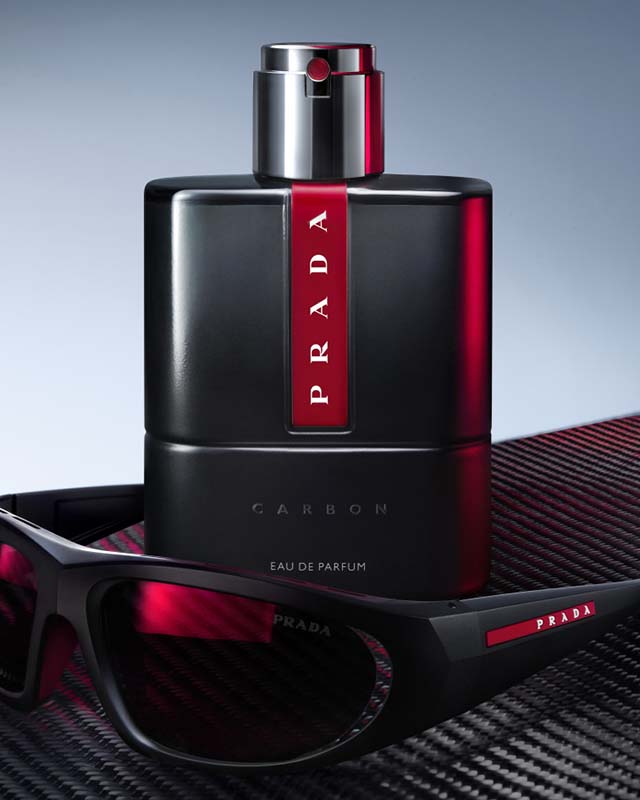 A Prada Carbon perfume bottle and sunglasses with red accents on a textured surface.