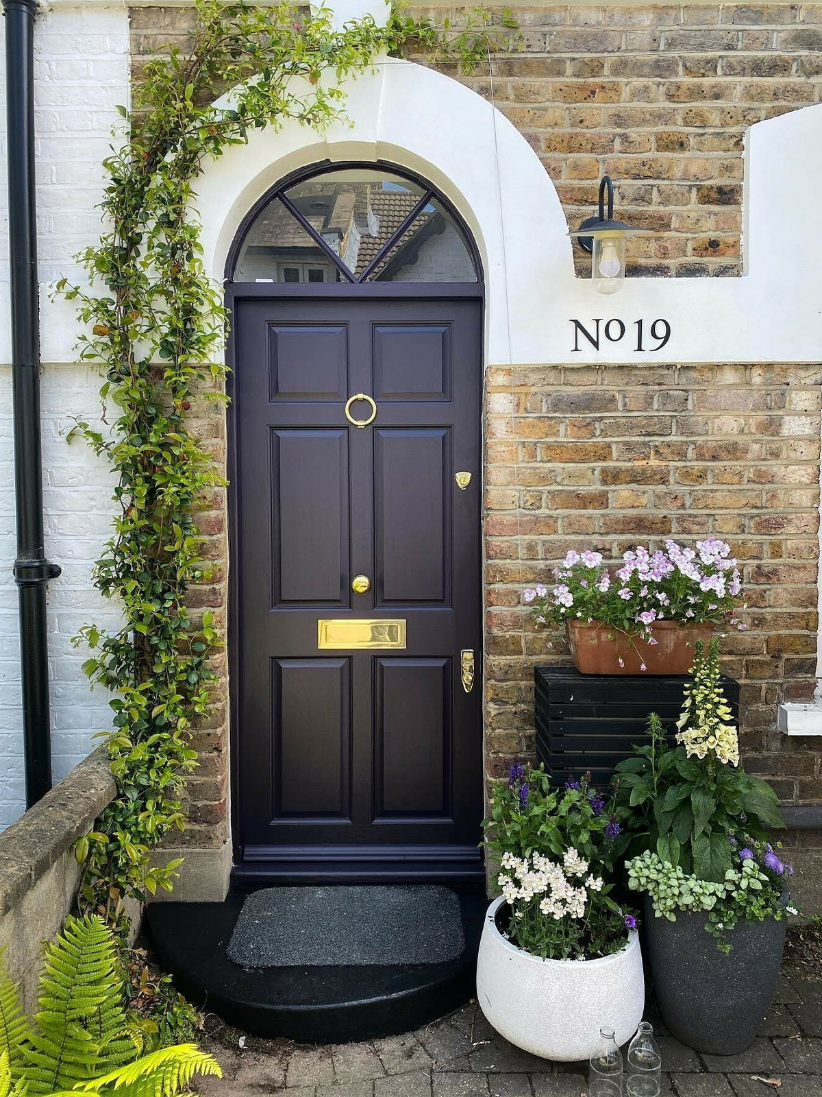 A dark navy front door on a brick house, framed by white masonry and surrounded by potted plants and climbing greenery.