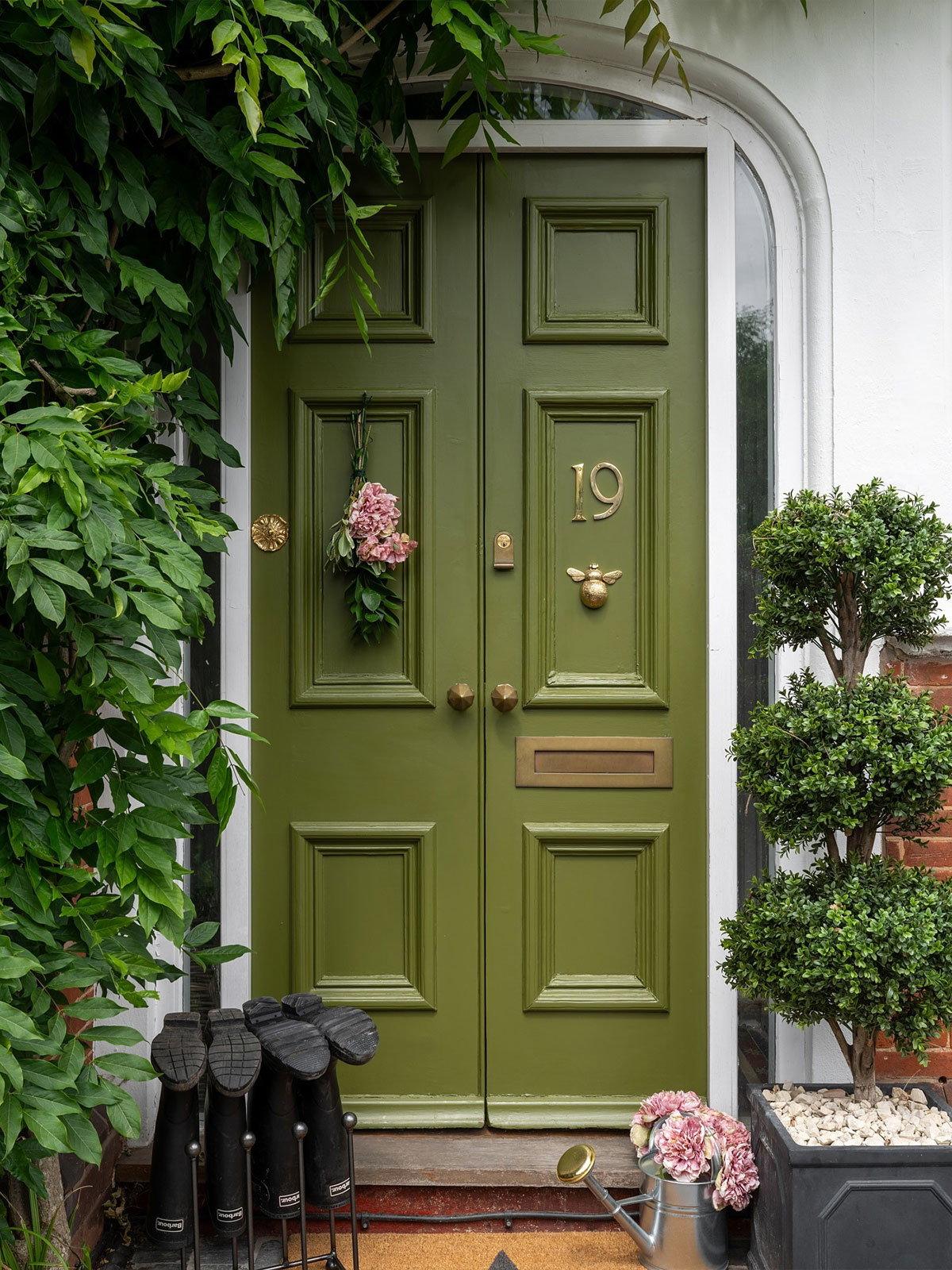 A neat, olive‑green double front door with brass details, pink flowers and leafy plants arranged around the entrance.