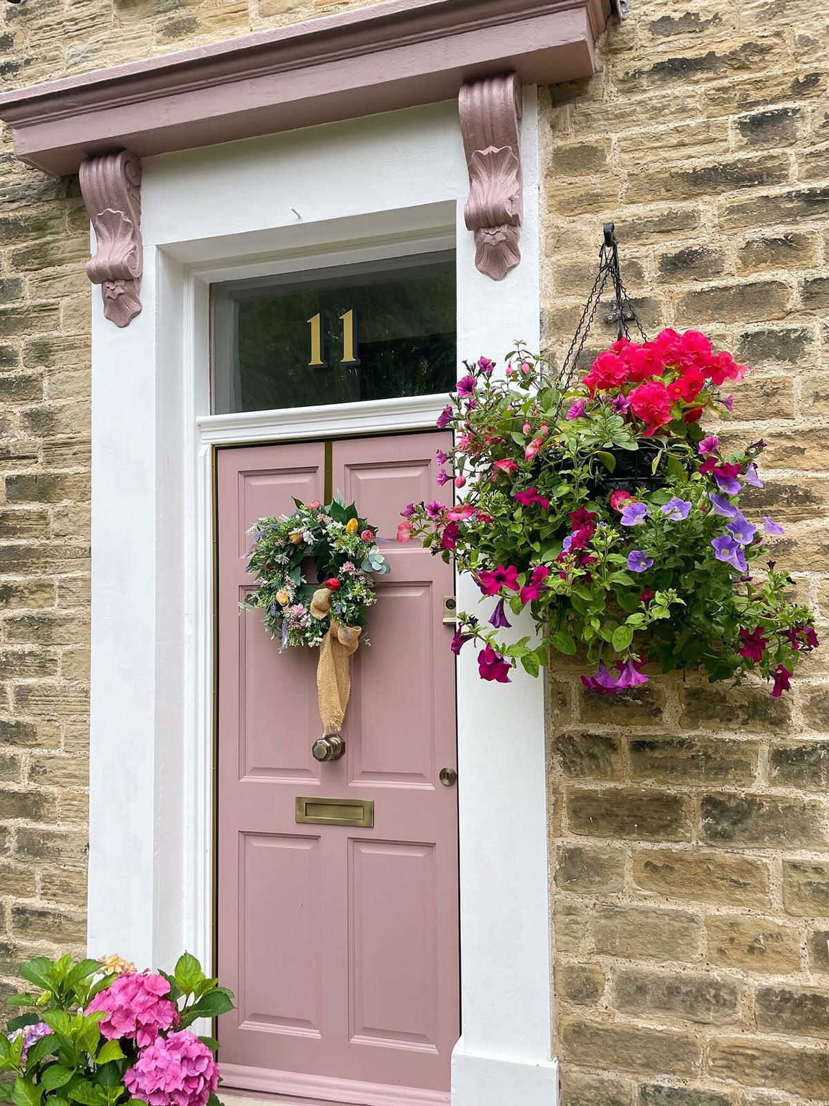 A soft pink front door on a stone house, decorated with a floral wreath and surrounded by bright flowers.