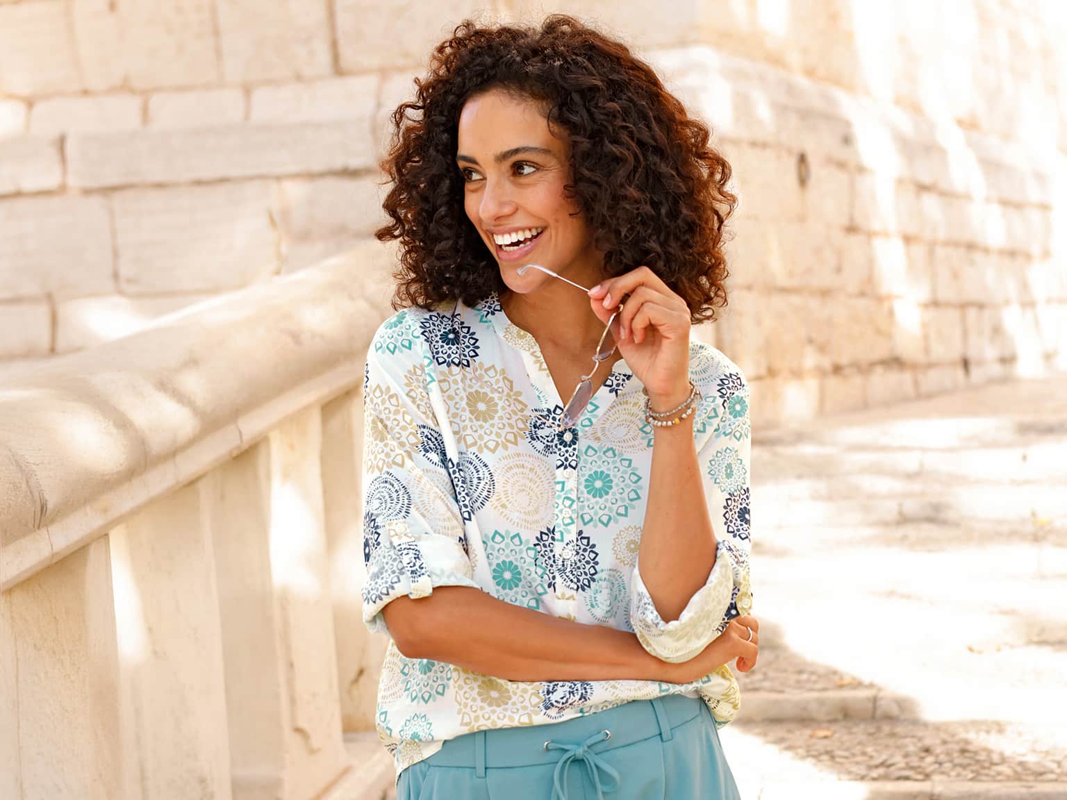 Ein Model trägt eine helle Bluse mit buntem Mandala-Muster und hochgekrempelten Ärmeln, sitzt an einer hellen Steinmauer im Freien.