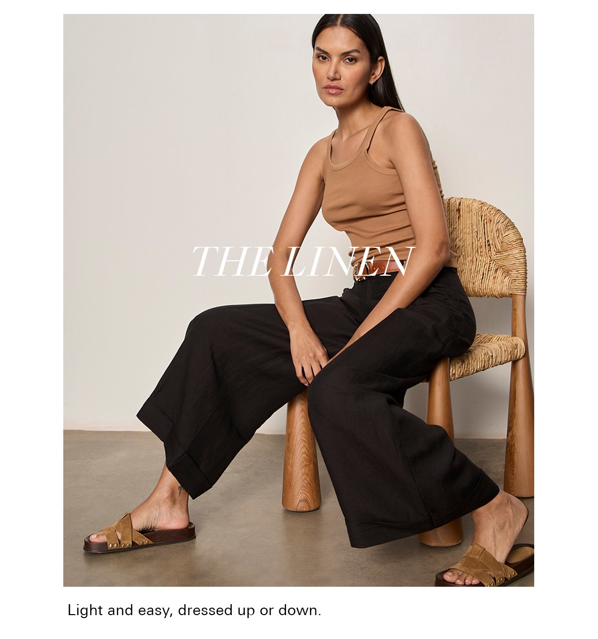 A woman sits on a wooden chair wearing a tan tank top, black wide-leg pants, and tan sandals. The text "THE LINEN" is overlayed on the image. The setting is minimal with neutral-colored walls and a concrete floor.