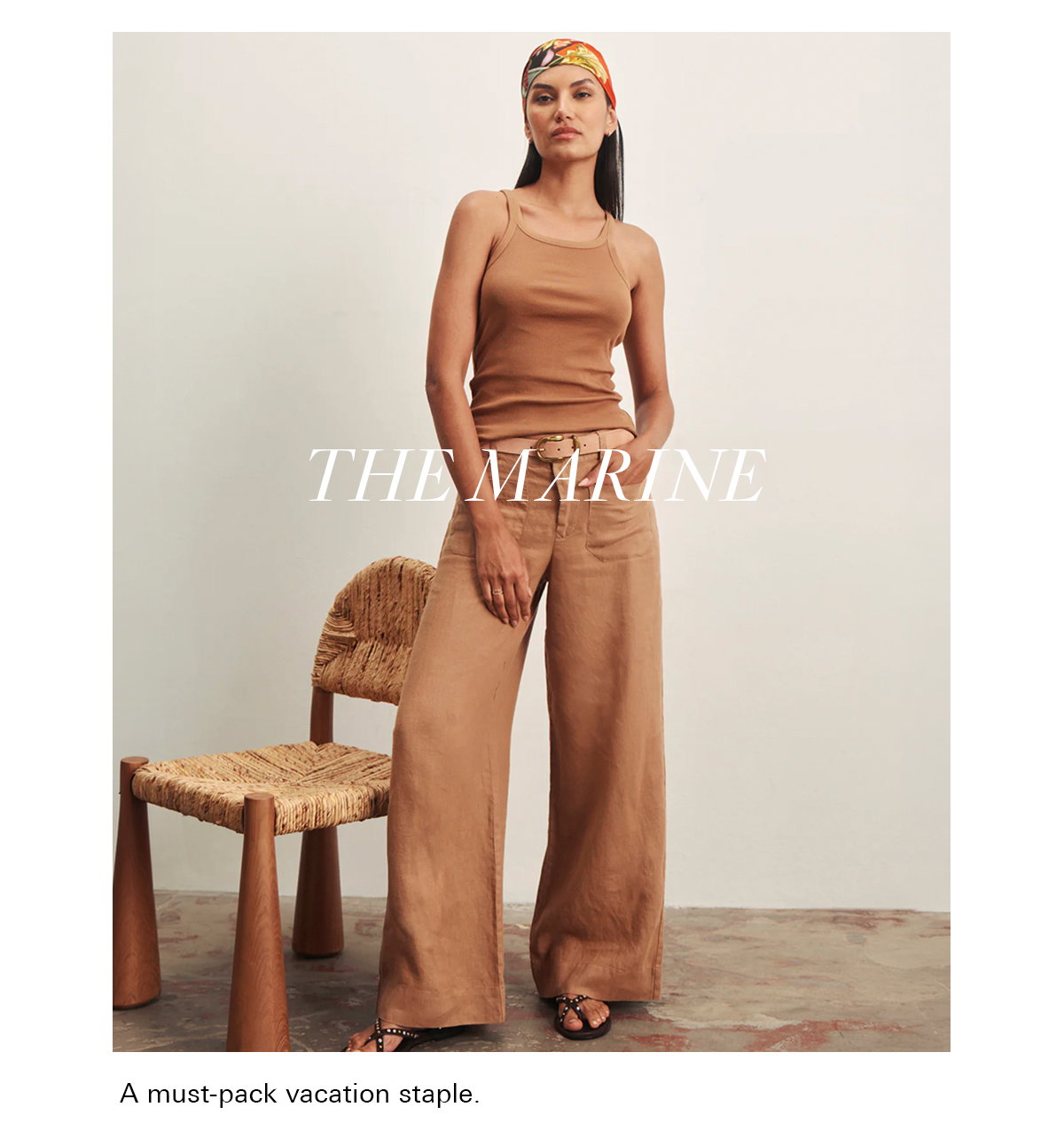 A woman stands indoors wearing a tan sleeveless top, wide-leg pants, sandals, and a colorful headscarf. She stands beside a woven chair on a concrete floor. Text reads, "THE MARINE." Caption: "A must-pack vacation staple.